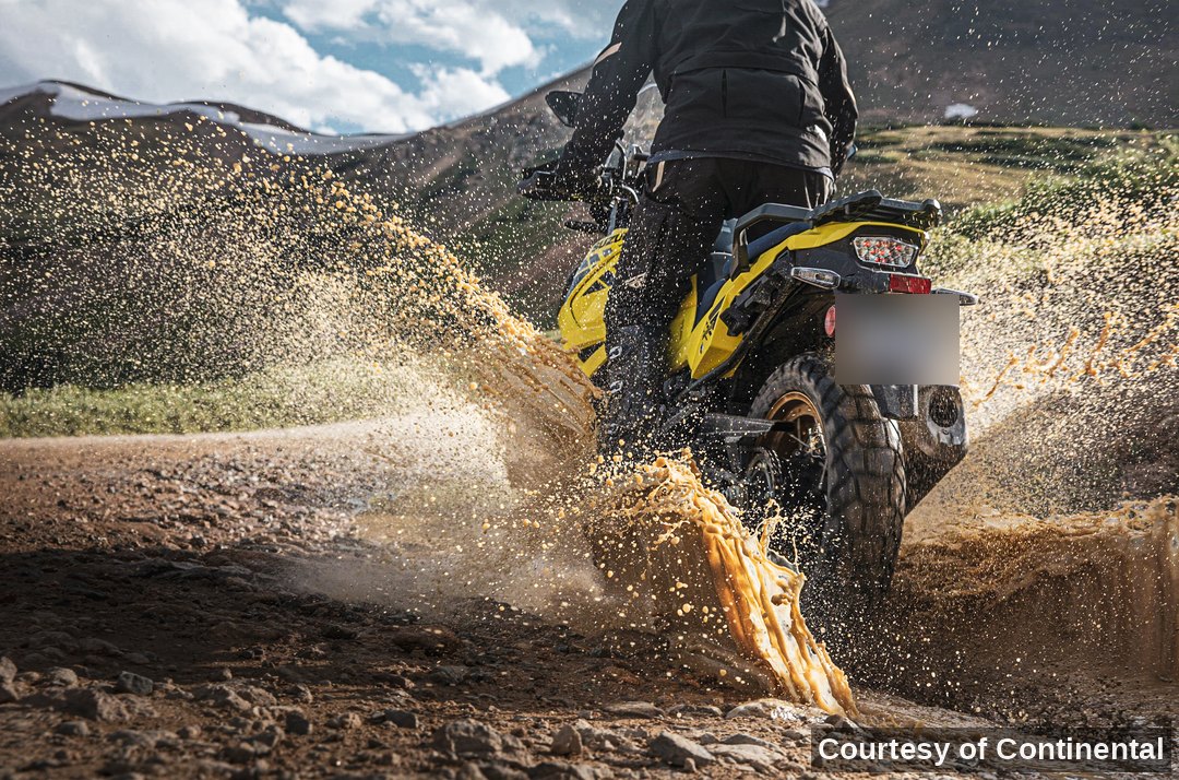 Rear view of a rider on a yellow adventure bike throwing a golden mud splash, mountains behind